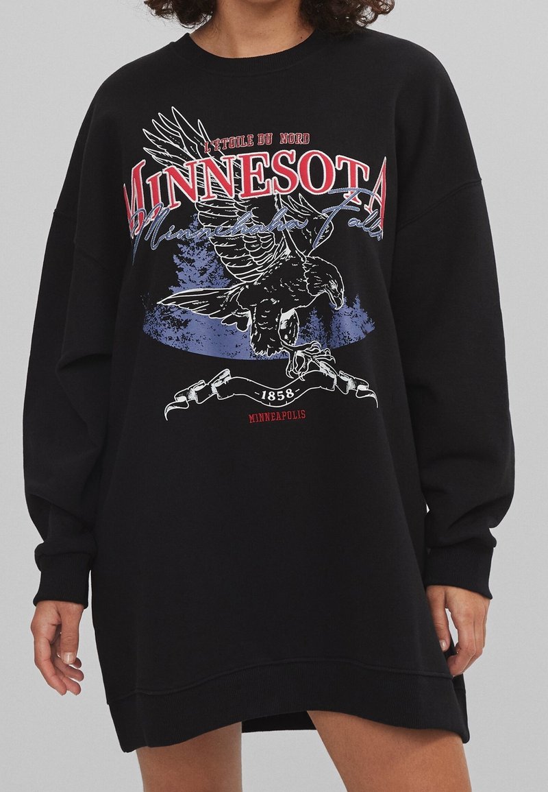 Personne portant un sweat-shirt noir oversize avec le texte "Minnesota", un graphisme d'aigle et des détails "Minneapolis 1858" à l'avant.