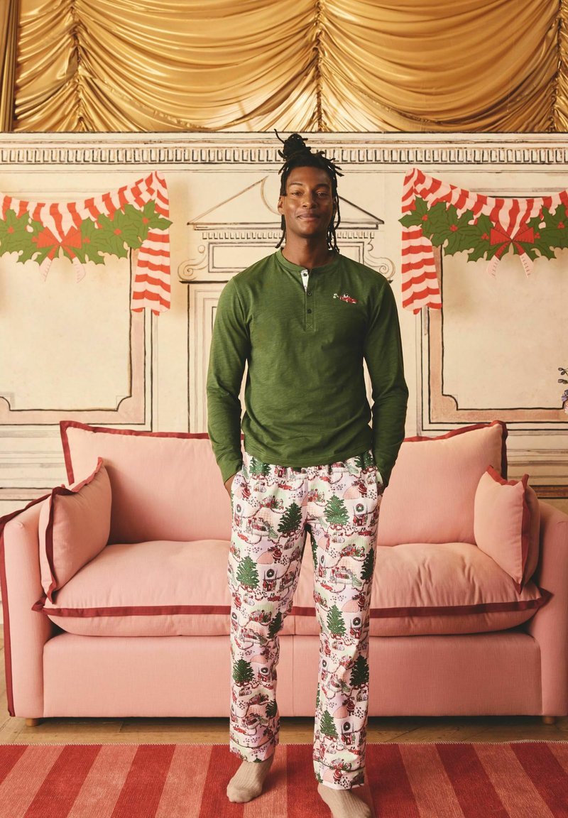 Grön långärmad henley med knappslå, i kombination med festligt mönstrade pyjamasbyxor med träd och julmotiv, stående på en rosa soffa.