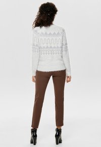 Vit, fluffig tröja med geometriskt mönster i ljusgrått, kombinerad med figurnära bruna byxor och svarta ankelstövlar. Enfärgad bakgrund.