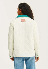 Prešívaná biela bunda s tyrkysovým golierom, so ružovou nášivkou na chrbte, bočnými vreckami so zelenými gombíkmi a voľným strihom.