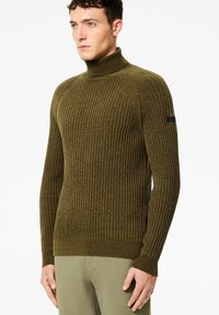 Uomo che indossa un maglione a collo alto a coste verde oliva e pantaloni leggeri verde oliva, in piedi con le mani rilassate sui fianchi, che guarda a sinistra.