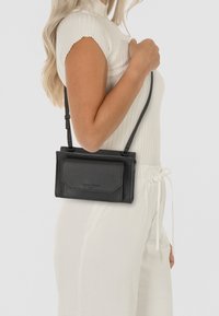 Czarna skórzana torebka na ramię z teksturowaną powierzchnią, prostokątnym kształtem, regulowanym paskiem oraz przednią kieszenią z klapą. Widoczne logo marki.