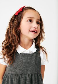 Graues Kleid mit einem gerafften Rock und Knopfdetails, getragen über einem weißen Hemd mit Kragen. Welliges braunes Haar mit einer roten Schleife.