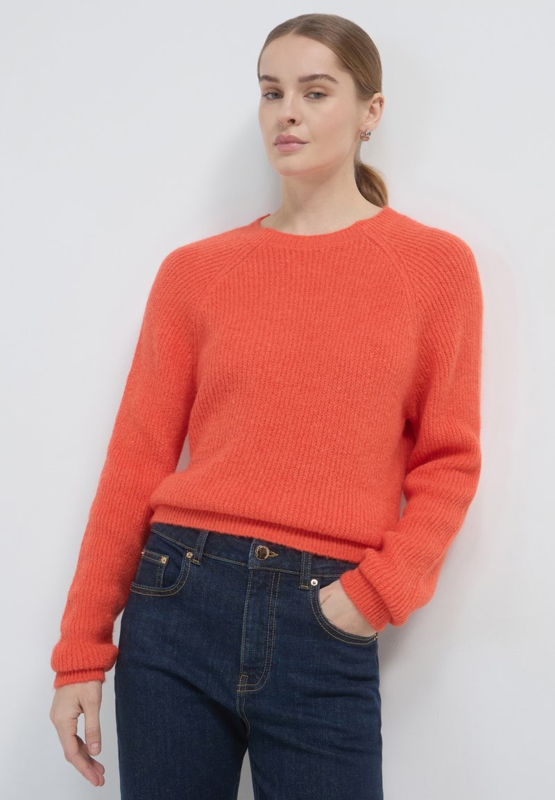 Orangener Strickpullover mit strukturierter Oberfläche und gerippten Bündchen, kombiniert mit dunkelblauen Jeans. Verfügt über einen runden Ausschnitt und einen lockeren Schnitt.