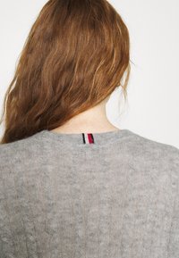 Pull en maille torsadée gris avec un col rond, présentant un motif texturé subtil et un accent rayé coloré à l'arrière du cou.