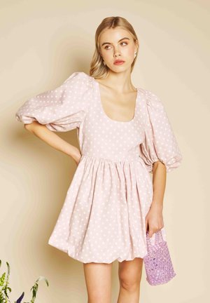 Femeie purtând o rochie roz deschis cu buline și mâneci bufante, ținând o geantă lavandă cu mărgele, stând în fața unui fundal bej.