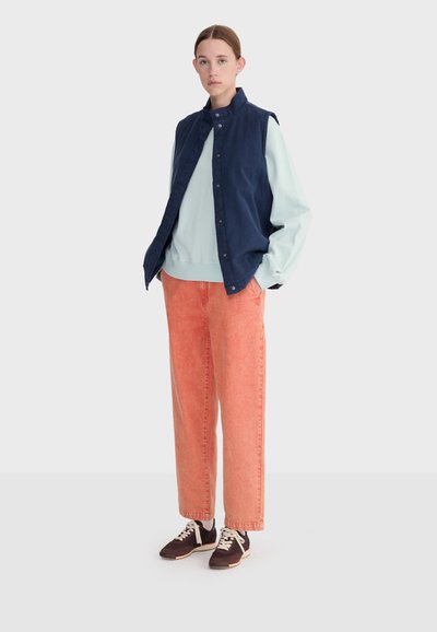 Jeune femme aux cheveux attachés portant un gilet sans manches bleu marine, un sweat-shirt bleu clair, un pantalon large corail et des baskets marron, debout les mains dans les poches.