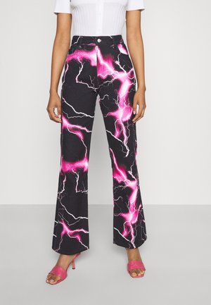 Personne portant un pantalon noir évasé avec des motifs d'éclairs roses vifs et blancs, associé à des sandales roses à talons et un haut blanc à manches courtes.