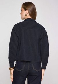 Giacca nera con sottili righe, dal design corto, dotata di un polsino con bottone, texture morbida e dettaglio alla schiena. Abbinata a jeans scuri.