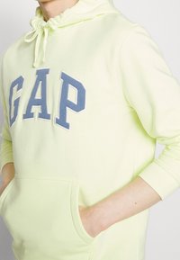 Lichtgele hoodie met een groot blauw "GAP" logo op de voorkant, een trekkoordcapuchon en een kangoeroezak. Zacht materiaal en geribde manchetten.