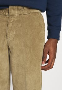 Bruna kordbyxor med mjuk textur, med rak passform och ribbat mönster. En hand syns på sidan, nära midjan.