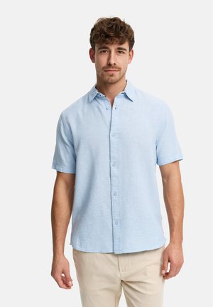 Mężczyzna stoi na jednolitym tle, ubrany w jasnoniebieską koszulę z krótkim rękawem zapinaną na guziki oraz beżowe spodnie, zwrócony przodem z neutralnym wyrazem twarzy.