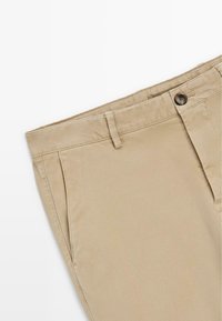 Pantalon en coton beige avec poches avant, passants de ceinture et fermeture à bouton sur fond blanc.