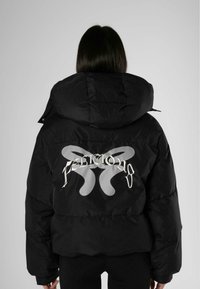 Zwarte pufferjack met een groot zilveren logo op de achterkant, voorzien van een strakke capuchon en een boxy silhouet. Gladde, glanzende textuur.