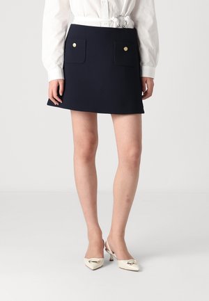 Kvinde iført mørkeblå mini nederdel med forlommer og guldknapper, hvid bundet bluse og cremefarvede lave spidse sko.