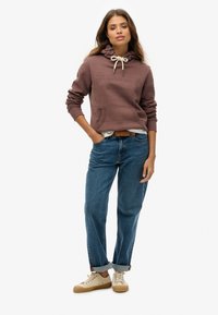 Sudadera con capucha marrón, con un bolsillo frontal y cordón, combinada con jeans claros remangados y zapatillas beige.