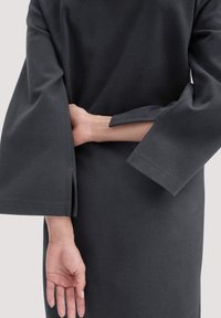 Dunkelgraues Kleidungsstück mit weiten, gespaltenen Ärmeln. Der Stoff wirkt weich und hat eine glatte Textur. Eine Hand hält den gegenüberliegenden Arm hinten.