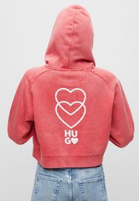 Sudadera recortada roja con capucha, que presenta un gráfico blanco de tres corazones superpuestos y la palabra "HUG" en la parte posterior. Textura suave.