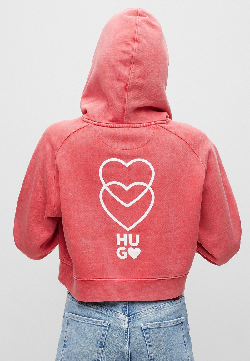 Sudadera recortada roja con capucha, que presenta un gráfico blanco de tres corazones superpuestos y la palabra "HUG" en la parte posterior. Textura suave.