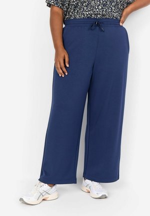 Marineblauwe brede sweatpants van zacht materiaal, met een trekkoord in de taille en platte naden voor extra comfort. Gekleed met witte sportschoenen.