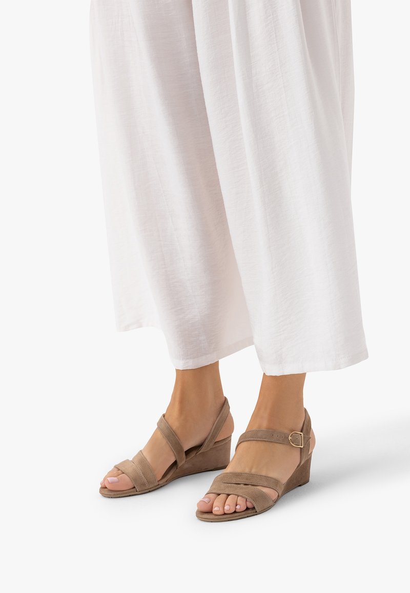 Stopy w beżowych zamszowych sandałach na koturnie z paskami wokół kostki i białe spodnie o szerokich nogawkach.