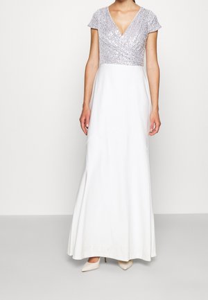 Femme portant une robe blanche longue jusqu'au sol avec un corsage en V à sequins argentés et des manches courtes, associée à des talons blancs pointus.