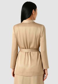 Blazer beige su misura con maniche lunghe e vita aderente. Tessuto liscio, colletto con rever e design a strati che si estende fino a metà coscia.