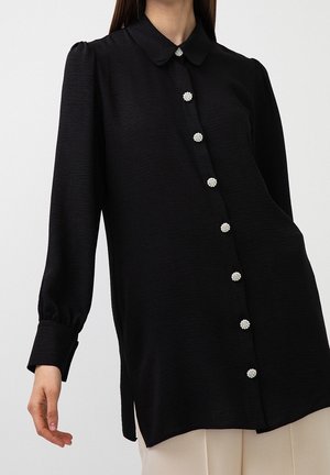 Sort langærmet bluse med puffede skuldre og knapper i perleklynger foran, båret med beige bukser mod en ensfarvet baggrund.