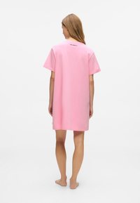 Femeie în picioare desculţă, întoarsă cu spatele, purtând o rochie scurtă roz, cu mânecă scurtă, având un mic logo pe partea superioară a spatelui.