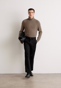 Brun strikket turtleneck sweater, sorte skræddersyede bukser og sort lædertaske; model der går mod en neutral baggrund.