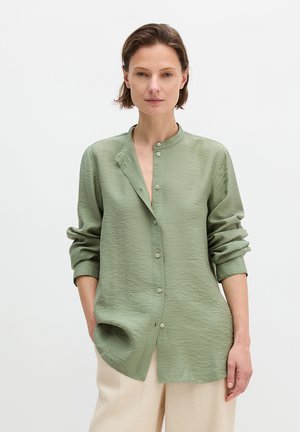 Kobieta ubrana w luźną, fakturowaną koszulę w kolorze szałwiowym z chińskim kołnierzykiem oraz kremowe szerokie spodnie, stojąca na jednolitym tle.