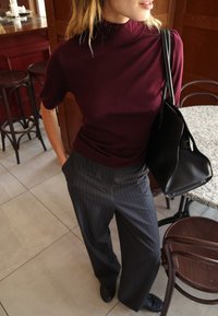 Burgundia värvi kinnine kaelusega top, mis on kombineeritud hallide peene triibuga laiade pükstega. Must käekott, millel on struktureeritud disain, toetub puusale.