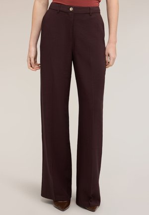 Femme portant un pantalon large marron foncé avec un pli devant, fermeture par boutons, poches latérales, assorti à des chaussures marron pointues et un haut orange rouille.