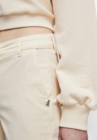 Beige kort tröja med ribbad mudd, kombinerad med matchande beiga byxor som har en dekorativ bi-charm och subtil märkning på ärmen.