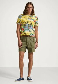 Ung man iklädd en gul blommig tropisk mönstrad t-shirt, olivgröna cargo-shorts och marinblå sneakers, stående mot en enkel vit bakgrund.