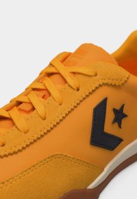 Κοντινό πλάνο ενός φωτεινού πορτοκαλί αθλητικού παπουτσιού με υφή σουεντ λεπτομερειών, κορδόνια στο ίδιο χρώμα και σκοτεινό αστέρι με λοξό λογότυπο στη πλευρά.