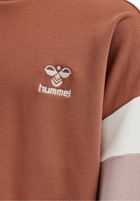 Hummel sweatshirt i rostfärg med beige och ljusrosa accenter. Har en broderad logotyp på bröstet, tillverkad av mjukt tyg.