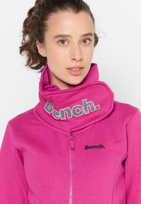 Fuchsiová mikina na zips s vysokým golierom a čiernym sieťovaným logom na golieri a menším logom na hrudi. Mäkka, štruktúrovaná tkanina.