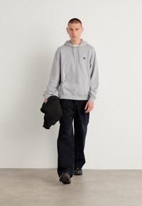 Grijze hoodie met capuchon en voorzak, gecombineerd met zwarte loszittende broek. Model houdt een zwarte jas vast; minimalistisch ontwerp op een neutrale achtergrond.