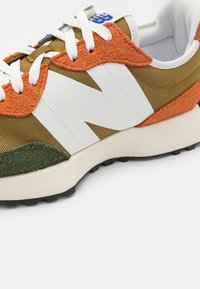 Detailní záběr na hnědou a oranžovou tenisku s bílými tkaničkami, velkým bílým logem "N", zeleným semišovým akcentem a černou podrážkou.