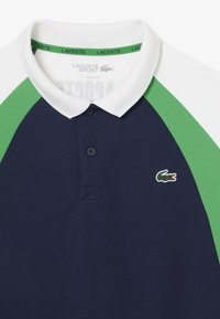 Polo navy con colletto bianco e dettagli verdi. Presenta una patta con bottoni e un logo ricamato sul lato sinistro del petto. Tessuto testurizzato.