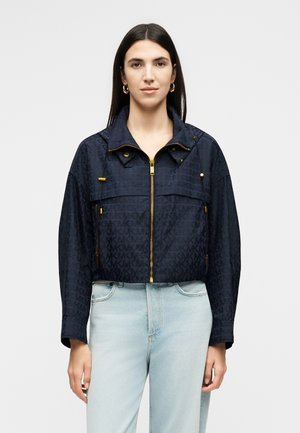 Kvinde med glat mørkt hår iført en navyblå cropped jakke med guldfarvet lynlås og lyseblå højtaljede jeans mod hvid baggrund.