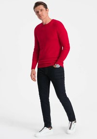 Chemise à manches longues en tricot rouge associée à un jean en denim foncé et des sneakers blanches avec des accents multicolores, ainsi qu'une montre ronde.