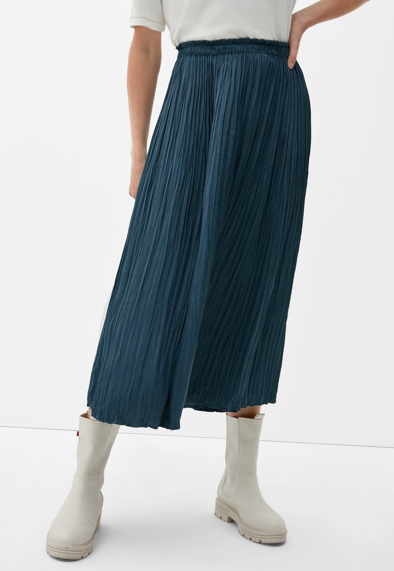 Personne portant une jupe midi plissée bleu marine associée à des bottines épaisses écrues sur un fond uni.