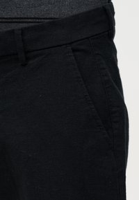 Gros plan sur une poche de pantalon noire et une boucle de ceinture portées avec une chemise texturée gris foncé rentrée à l'intérieur.