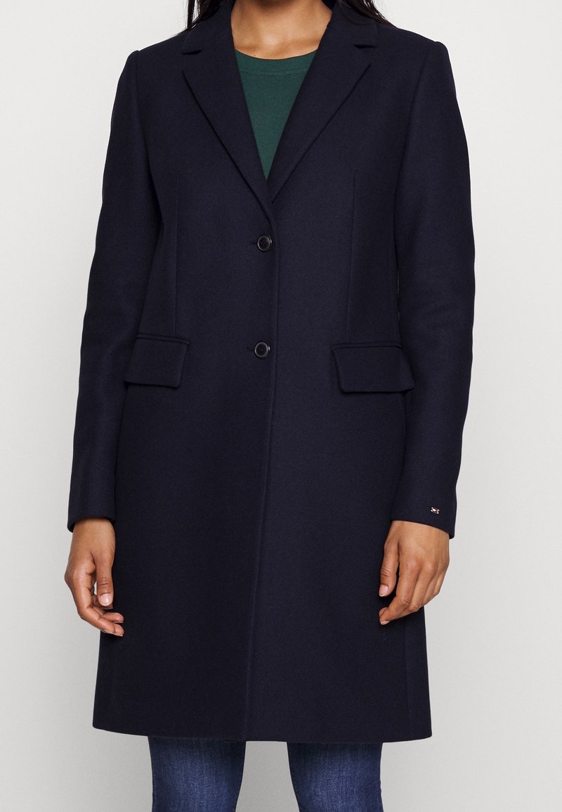 Cappotto lungo in misto lana di colore blu navy con rever a punta, due bottoni e tasche laterali. Presenta una superficie liscia e una vestibilità su misura.