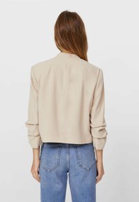 Blazer beige corto con maniche lunghe, vestibilità morbida e texture sottile. Abbinato a jeans blu a vita alta, mostrando la vista da dietro.