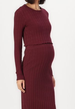 Femme enceinte portant un haut bordeaux moulant à manches longues côtelé et une jupe assortie, debout devant un fond blanc uni.