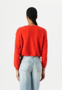 Pull rouge en peluche, court avec des manches longues, associé à un jean taille haute bleu clair. La vue de dos met en valeur la texture du pull.
