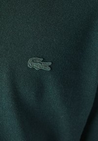 Tessuto a maglia di un verde scuro con una finitura testurizzata, caratterizzato da un logo ricamato che assomiglia a un coccodrillo sul lato sinistro.